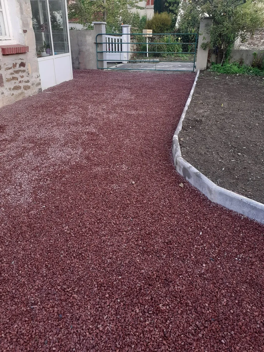 Allée en gravillon rouge entre deux bordures béton, le long d'une maison
